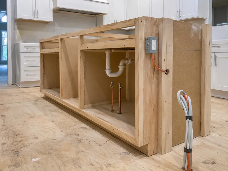 Kitchen island roughed in before the countertop, with a PVC drain and vent riser, copper supply lines on quarter-turn shutoffs, and an outlet box wired through the end panel
